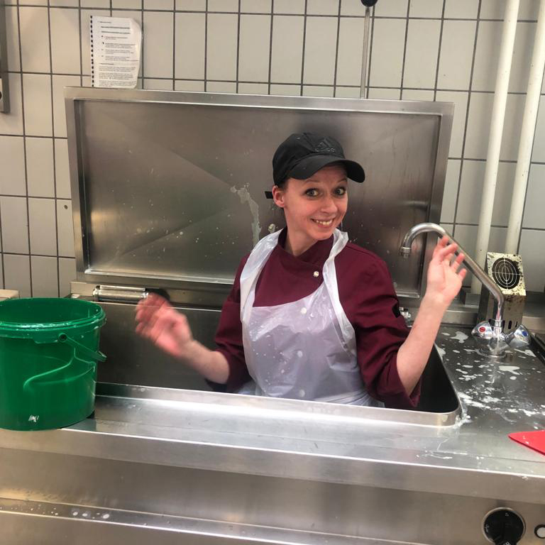 Eine lächelnde Frau in rotem Shirt und Kappe sitzt in einer Industriel-Spüle. Ein grüner Eimer steht daneben.