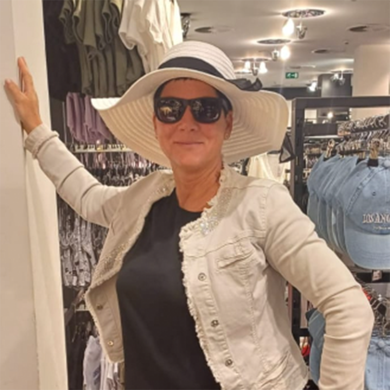 Frau mit weißem Hut und Sonnenbrille, lächelt freundlich, trägt beige Jacke in einem Geschäft.