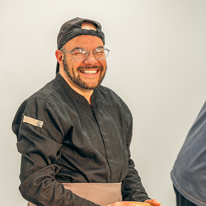 Ein lächelnder Mann mit Brille und dunkler Kochjacke trägt eine schwarze Kappe rückwärts